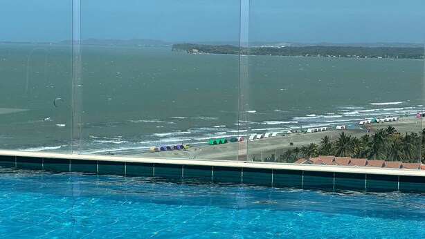 A pool with clear blue water, a beach with colorful tents, and a view of the ocean and distant hills. - Sunno Beach Condominium - Playa Azul (Cartagena)