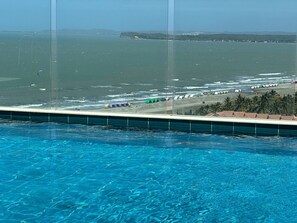 A pool with clear blue water, a beach with colorful tents, and a view of the ocean and distant hills. - Sunno Beach Condominium - Playa Azul (Cartagena)