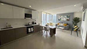 A modern kitchen with a dining area, a grey sofa, and a large window with a city view. - Sunno Beach Condominium - Playa Azul (Cartagena)