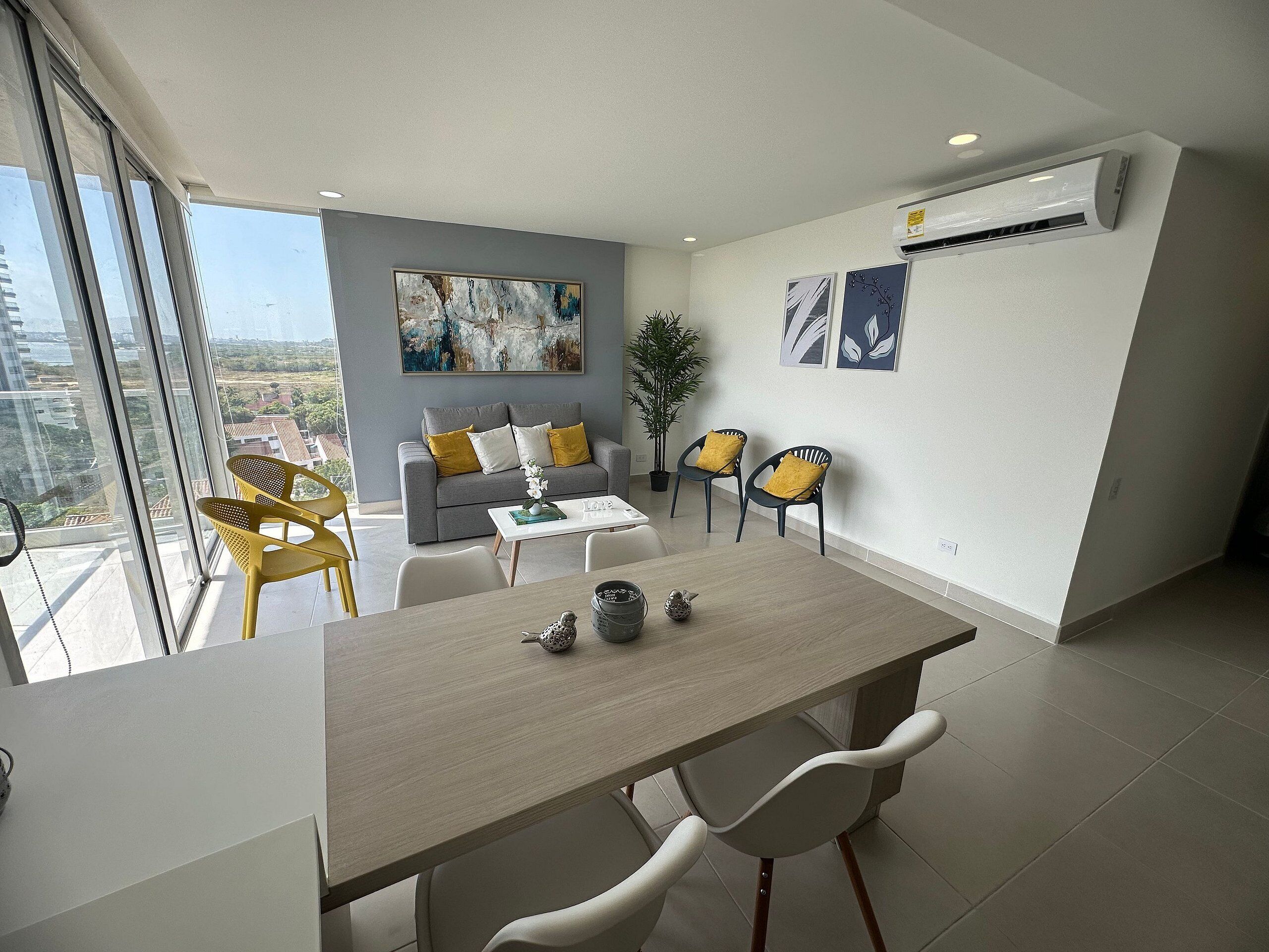 Una sala de estar moderna con un sofá gris, sillas amarillas, una mesa de comedor y un amplio ventanal con vistas a la ciudad.