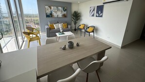A modern living room with a grey sofa, yellow chairs, a dining table, and a large window offering a city view. - Sunno Beach Condominium - Playa Azul (Cartagena)