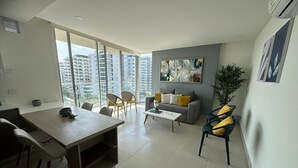 A modern living room with a dining area, large windows, a grey sofa, and a painting on the wall. - Sunno Beach Condominium - Playa Azul (Cartagena)