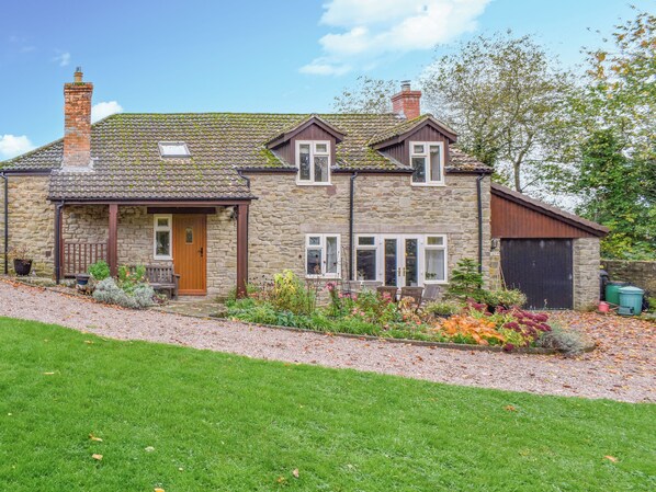 Une maison en pierre avec un toit en tuiles, un porche à l’avant et un jardin où poussent différentes plantes et fleurs.