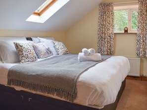 A neatly made bed with a grey blanket, white pillows, and a rolled towel on top. There is a window with curtains and a clock on the wall. - 3 bedroom accommodation in Lydney (Lydney)