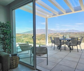 A modern patio with a glass door leading to a terrace. The terrace features a table set for dining with chairs, a potted plant, and a view of a mountainous landscape. - Penthouse with 2 large terraces and a Plunge Pool (La Cala de Mijas)