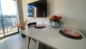Un comedor con una mesa blanca, sillas negras, un televisor de pantalla plana montado en la pared y un ventanal con vistas al verdor.