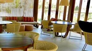 A spacious outdoor seating area with wicker furniture and a round table, featuring a view of greenery through large windows. - Apartment 2/4 Reserva Timeantube Praia do Forte - Comfort and Leisure in Nature (Açu da Torre)