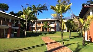 A courtyard with a paved walkway, palm trees, and a building with multiple balconies. - Apartment 2/4 Reserva Timeantube Praia do Forte - Comfort and Leisure in Nature (Açu da Torre)