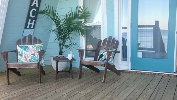 Una terraza de madera con dos sillas Adirondack, una planta en maceta y un letrero que dice 'BEACH Life'.