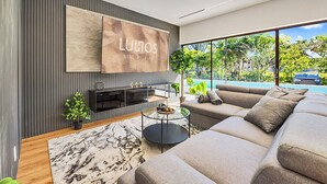 Una sala de estar moderna con un sofá grande, una mesa de centro y un televisor montado en la pared. Se ve una piscina a través de los amplios ventanales.