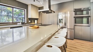 Una cocina moderna con una isla grande, electrodomésticos de acero inoxidable y vistas a árboles a través de las ventanas.