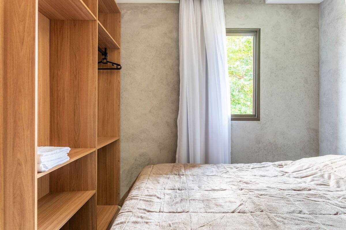 Une chambre avec une armoire en bois, un lit recouvert d’une couverture à motifs, une fenêtre donnant sur un paysage verdoyant et un rideau blanc.