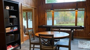 Una habitación con paneles de madera, una mesa redonda con sillas, una estantería y ventanas amplias con vistas a árboles.