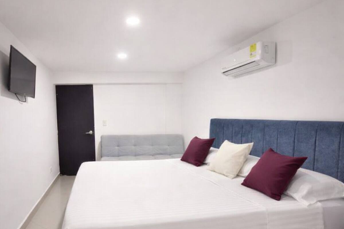 A bedroom with a bed, a grey headboard, a white mattress, and a wall-mounted air conditioner.