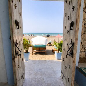 A wooden door leading to a beach view with a bed and lounge chairs.