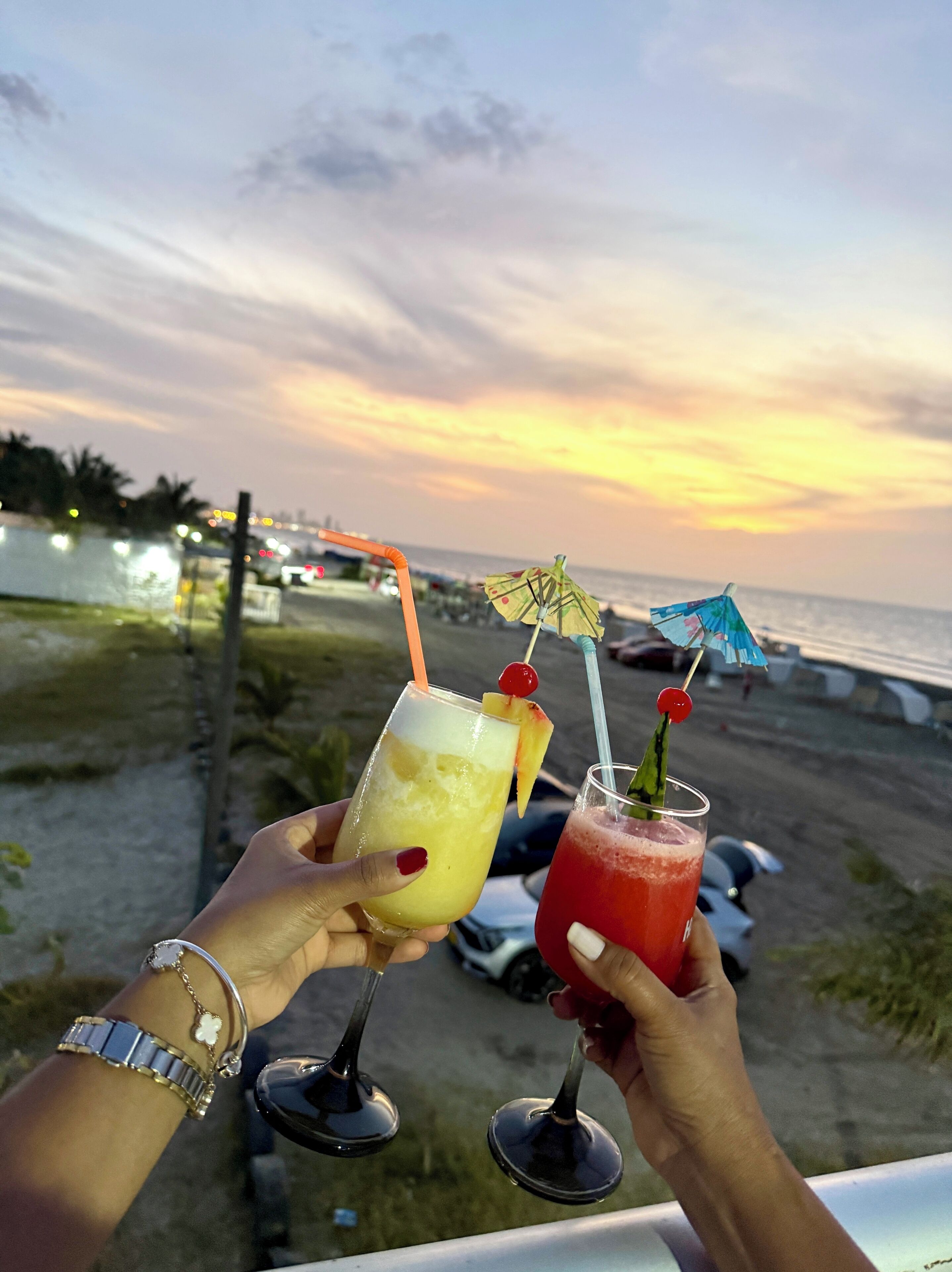 Zwei Hände halten Cocktailgläser mit Strohhalmen, eines mit einem gelben und das andere mit einem roten Getränk, vor einer Sonnenuntergangslandschaft.