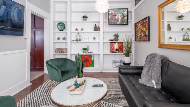 Un salón moderno con un sofá, un sillón verde, una mesa de centro y una estantería llena de plantas y objetos decorativos.