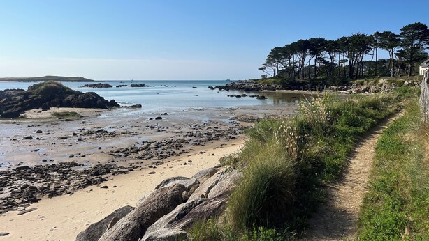 Eine Küstenlandschaft mit Sandstrand, Felsformationen und einem zu einem von Bäumen umgebenen Gebäude führenden Pfad.