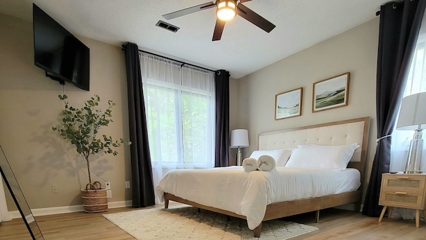 A bedroom with a bed, a ceiling fan, a TV, a plant, and a window with curtains.