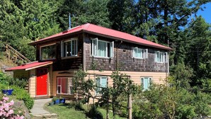 A two-story wooden house with a red roof, a front porch, and a stone pathway leading to the entrance. - Silva Bay View, a boutique, private BnB with a great view (Gabriola)