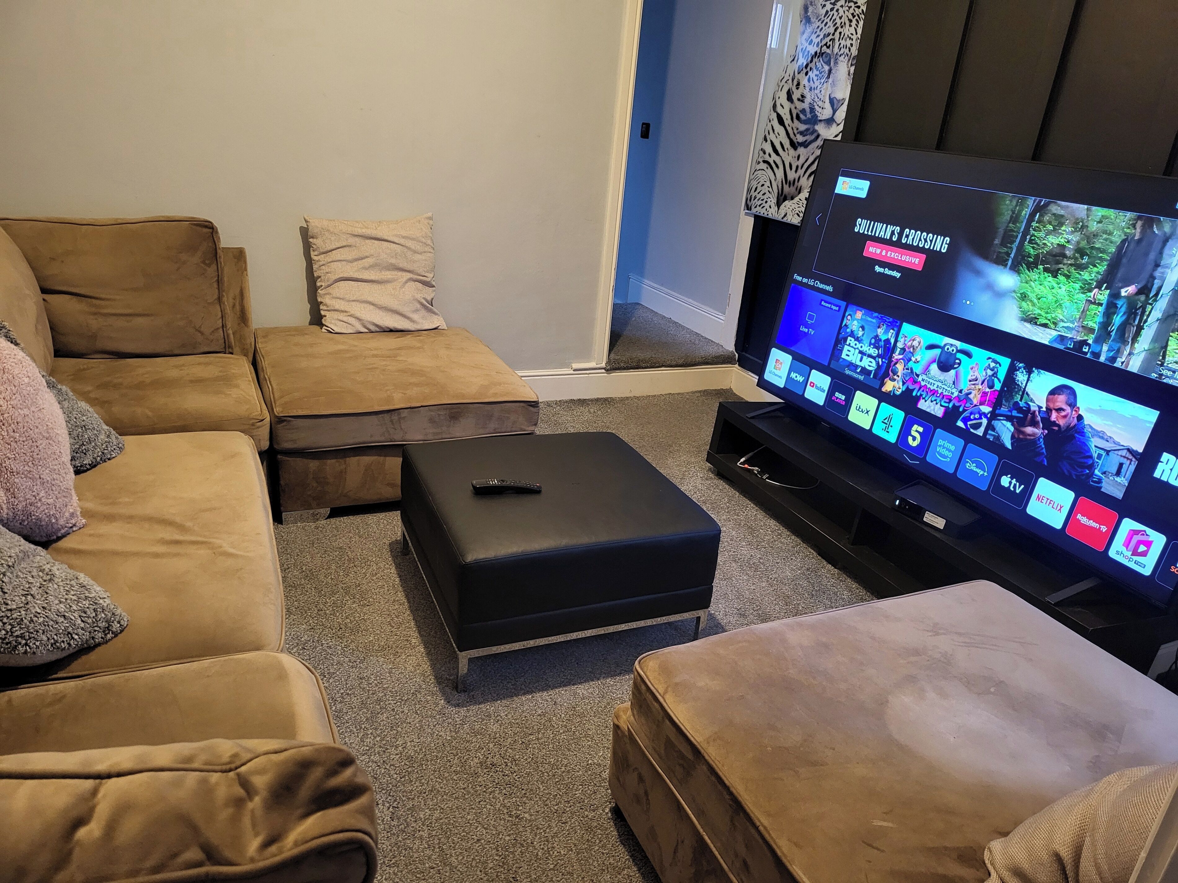 A living room with a large flat-screen TV displaying multiple channels, a brown sofa, a black ottoman, and a remote control on the ottoman.