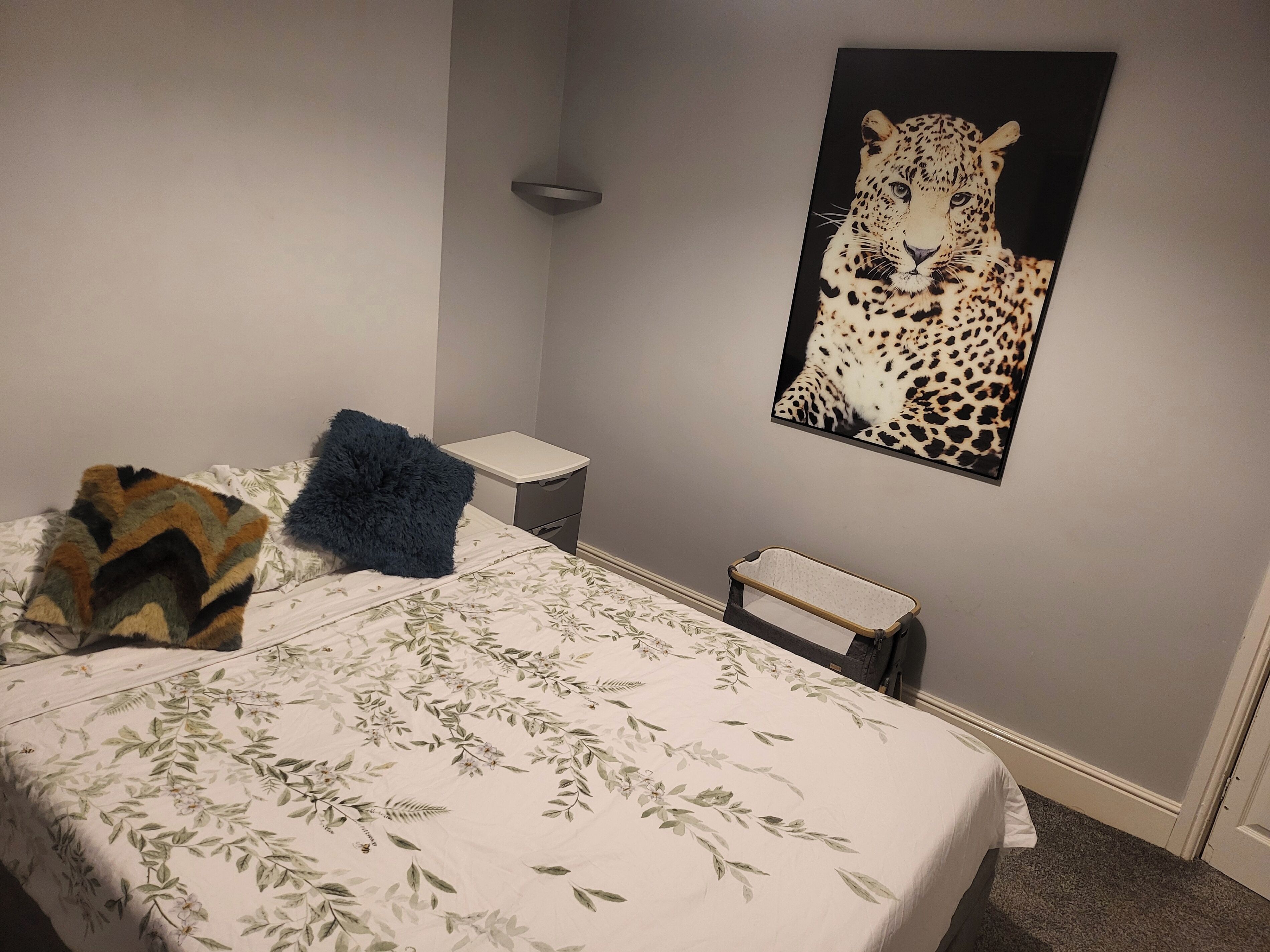 A bedroom with a bed, a nightstand, a small bedside table, a wall-mounted shelf, and a leopard print artwork.