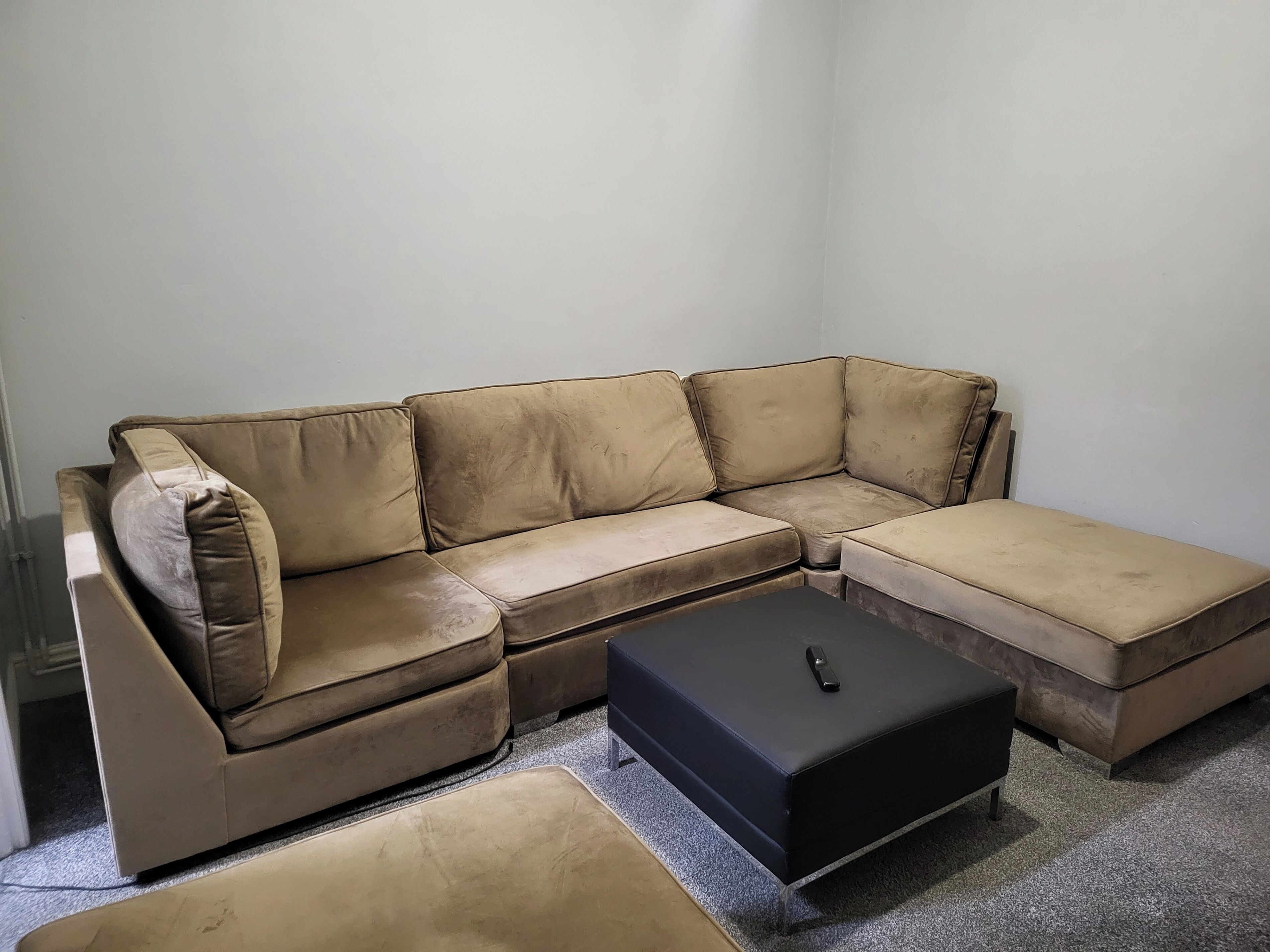 A beige sectional sofa with a black ottoman and matching ottoman, positioned against a plain wall.