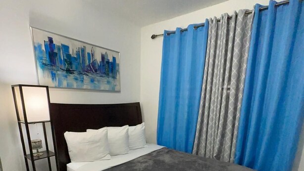 Una cama bien hecha con una manta gris, almohadas blancas y una toalla enrollada en el reposapiés. Un cuadro de un paisaje urbano adorna la pared, y cortinas azules cubren la ventana.