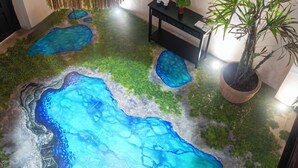 Een kamer met een uniek vloerontwerp dat een natuurlandschap nabootst, met blauwe en groene kleuren, een ronde spiegel aan de muur, een potplant en een kleine tafel met een lamp.