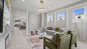 A modern living room with a sofa, ottomans, a coffee table, and a TV. There are windows, a door, and a chandelier.