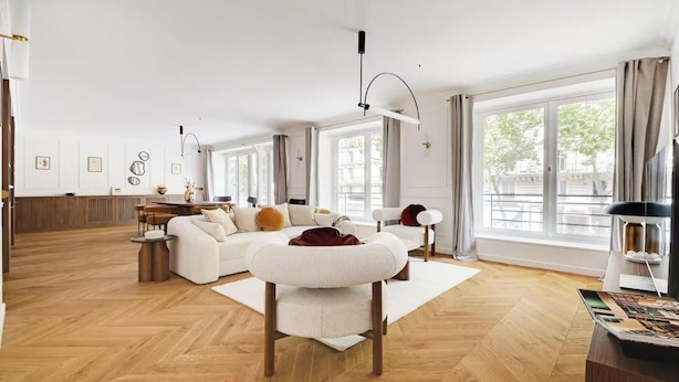 Ein geräumiges Wohnzimmer mit einer großen Couch, einem Couchtisch und einem Essbereich. Große Fenster sorgen für natürliches Licht und bieten einen Ausblick nach draußen.