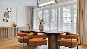 Ein Essbereich mit einem Holztisch, sechs orangenen Stühlen, einer Vase mit getrocknetem Pampasgras und Blick auf Gebäude durch große Fenster.
