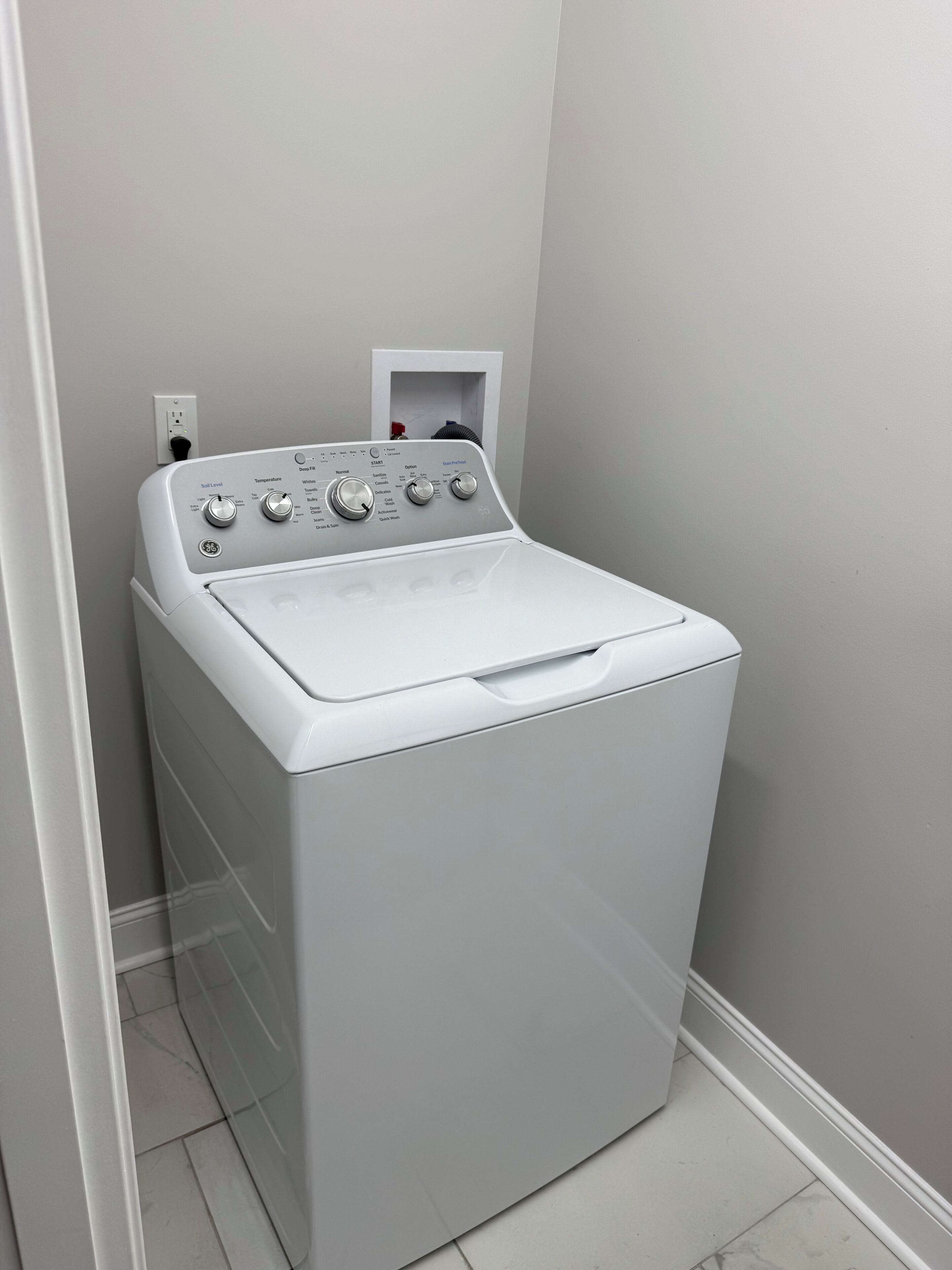 Eine weiße Waschmaschine mit Frontlader in einem Waschraum mit grauer Wand und weißen Fliesen.
