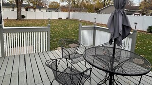 Een houten terras met een ronde metalen tafel en stoelen, een grijze parasol en een witte hekwerkhek.