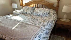 Een net opgemaakt bed met een bloemenbedekker, een sprei en twee kussens. Er staan twee nachtkastjes met lampen, een houten hoofdbord en een vloerkleed op de vloer.