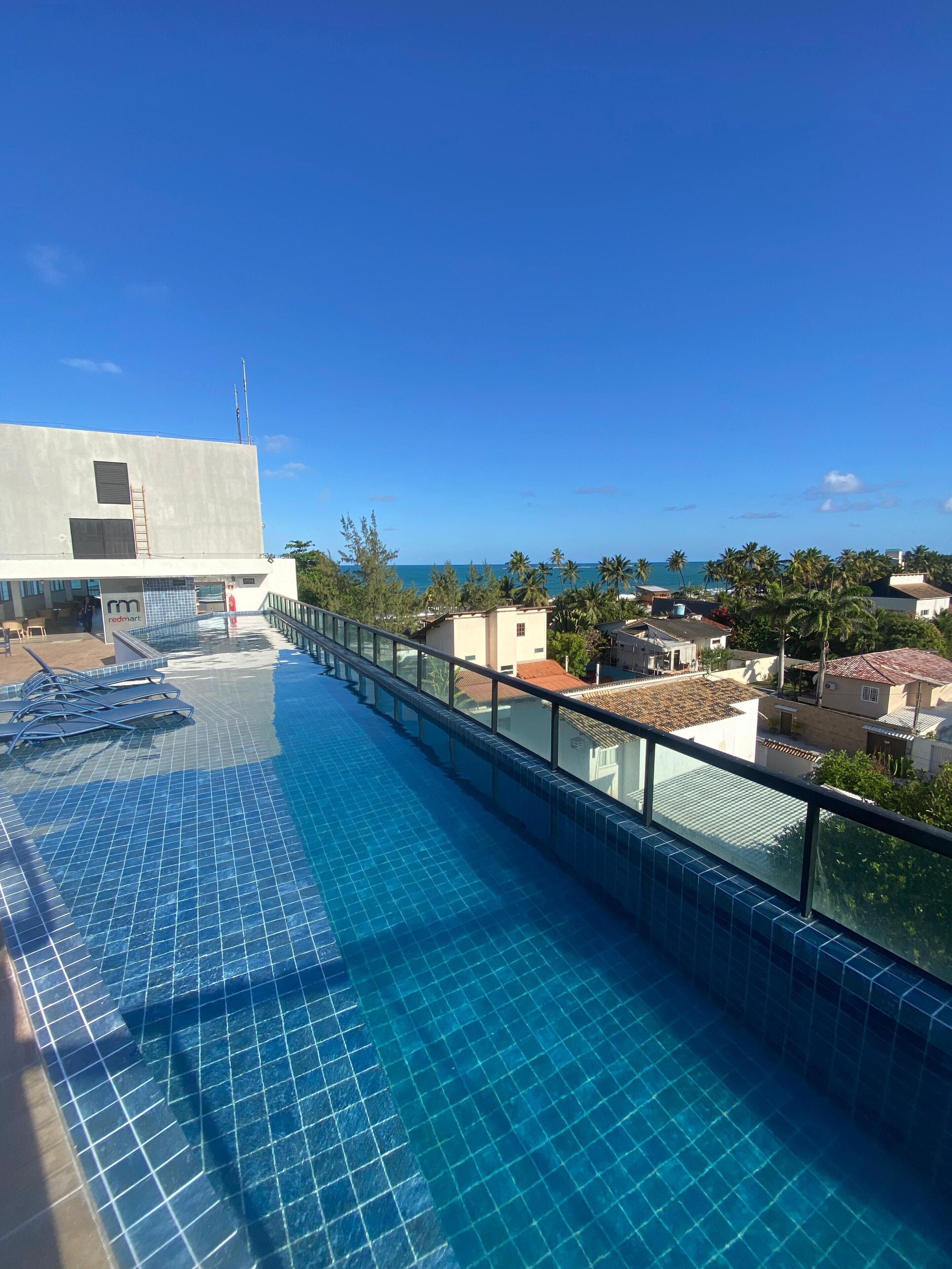 Una piscina al terrat amb aigua blava i cristal·lina, una barana de vidre i vista a l'oceà i els edificis circumdants.