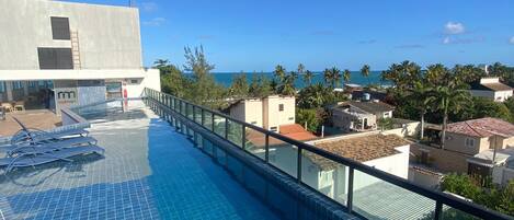 A rooftop pool with clear blue water, a glass balustrade, and a view of the ocean and surrounding buildings.