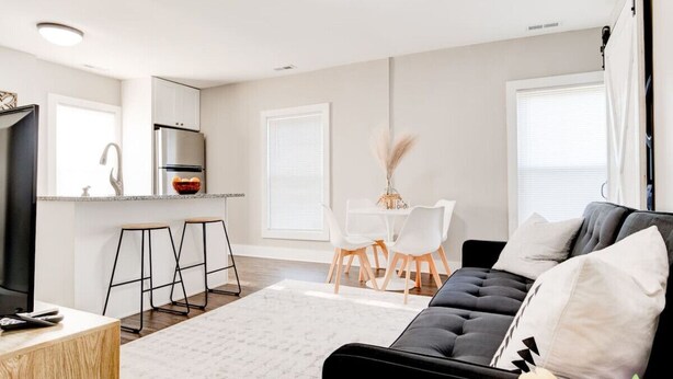 Una sala de estar moderna con un sofá negro, una mesa de comedor con sillas, una zona de cocina con refrigerador y un ventanal grande.