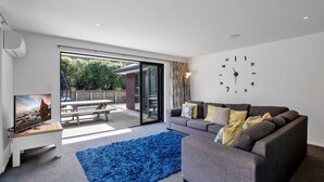 A modern living room with a grey sofa, a blue rug, a TV on a white stand, and a large window with a view of greenery.