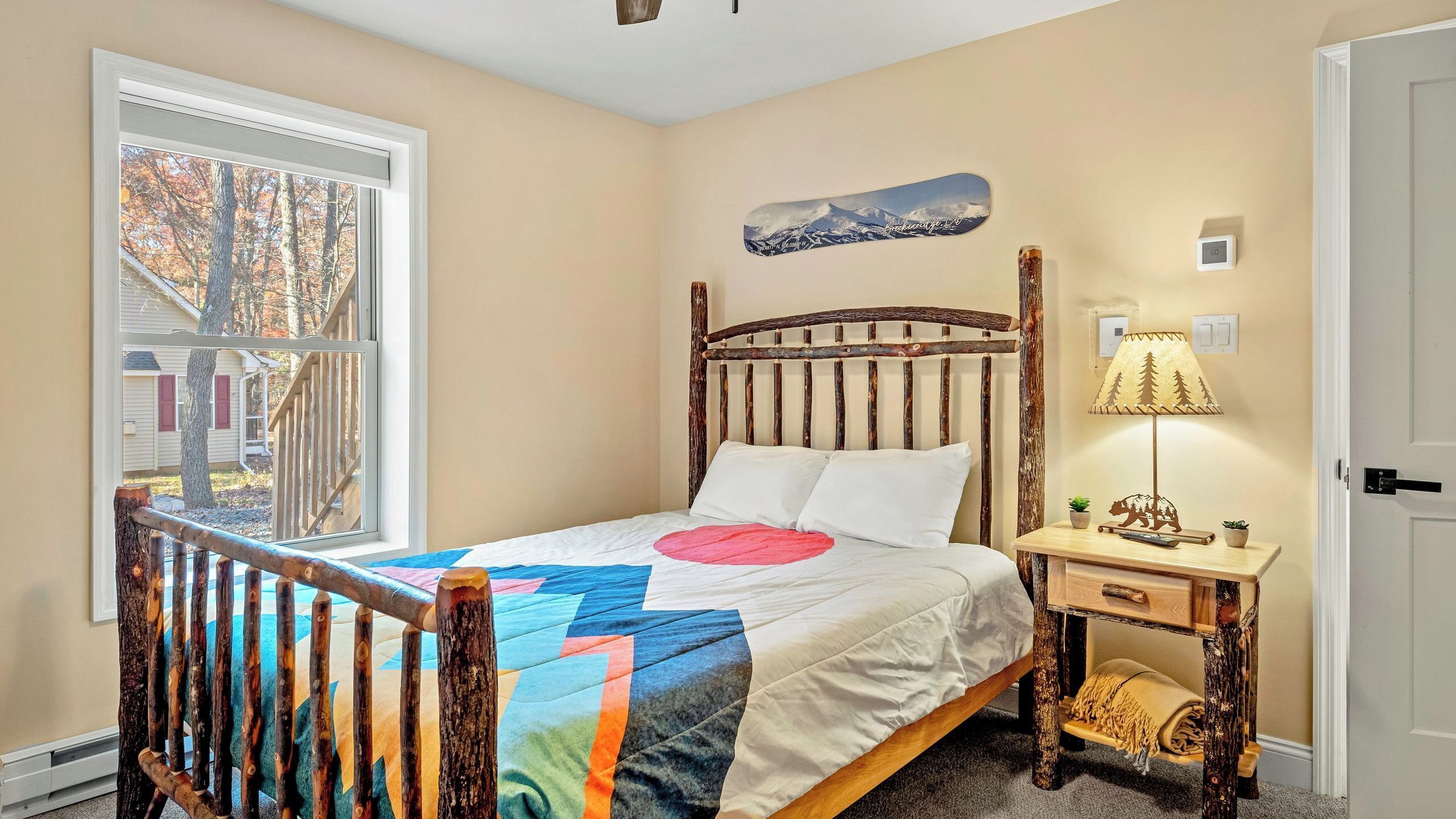 Een slaapkamer met een houten bed, een raam met uitzicht op bomen, een wandlamp en een klein nachtkastje.