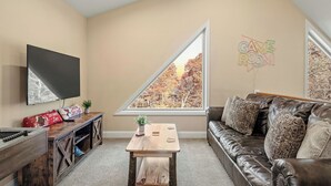 A living room with a TV, a wooden coffee table, a leather sofa, and a large window with a view of trees. - Stephen Way by Avantstay Hot Tub + Game Room Albrightsville (Albrightsville)