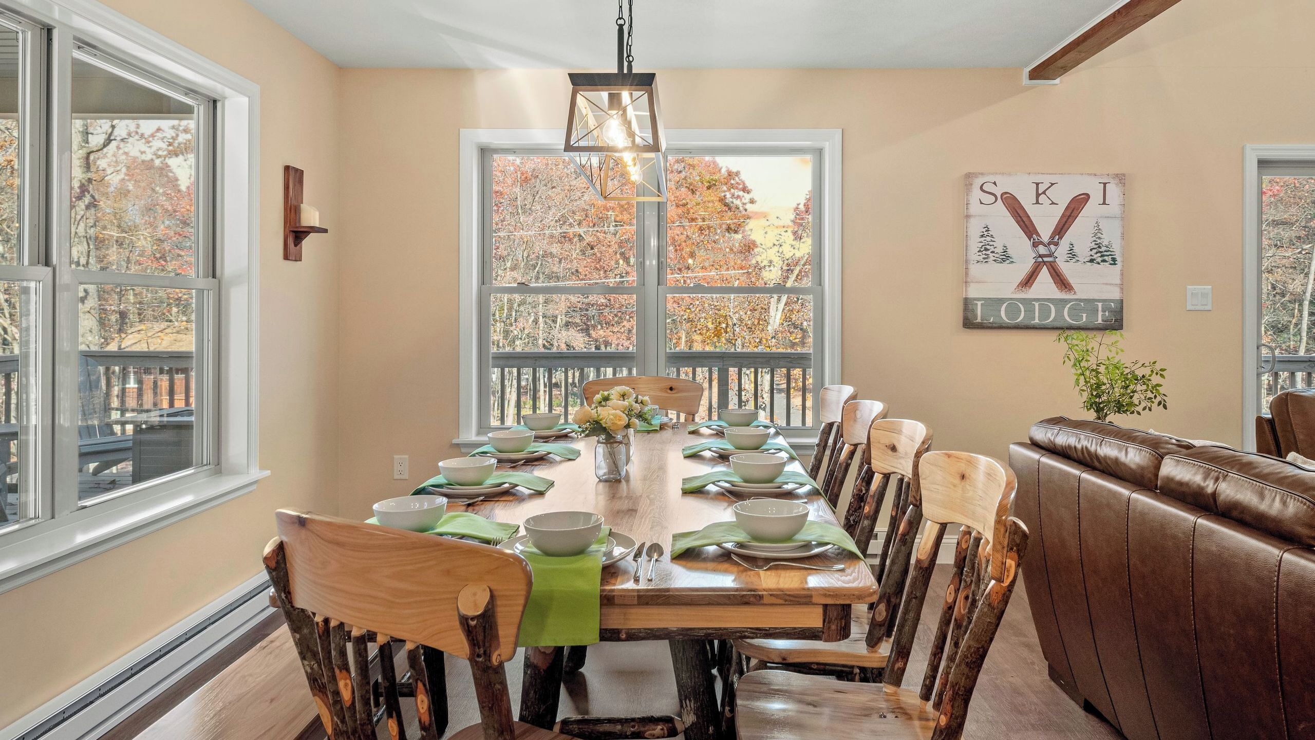 A dining room with a wooden table set for six, surrounded by wooden chairs, a brown leather couch, and a large window with autumn foliage view.