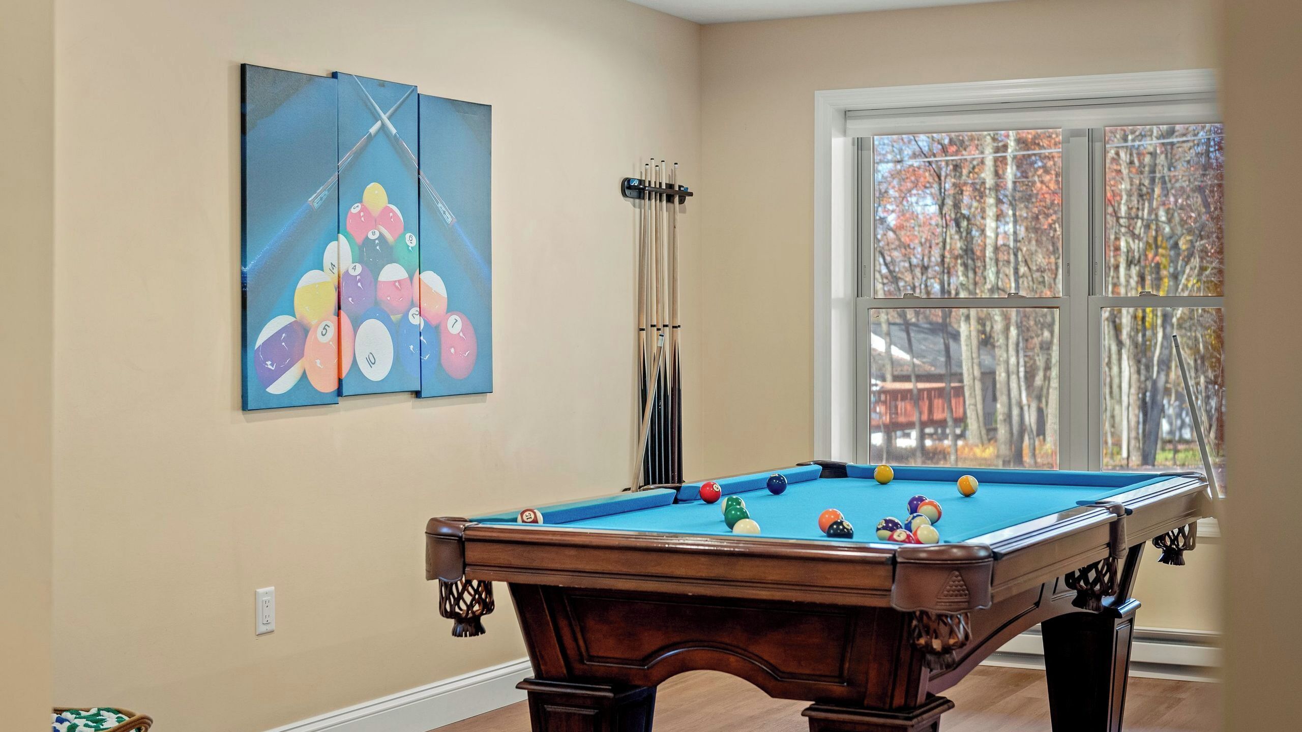 Una mesa de billar con bolas, una ventana que da a un paisaje de árboles y una pared con un soporte para tacos.