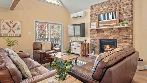 A living room with leather sofas, a stone fireplace, a TV, and a large window with a view of trees. - Stephen Way by Avantstay Hot Tub + Game Room Albrightsville (Albrightsville)
