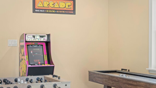 Una máquina de videojuegos retro, una mesa de futbolín y una mesa de billar en una sala con paredes color beige.