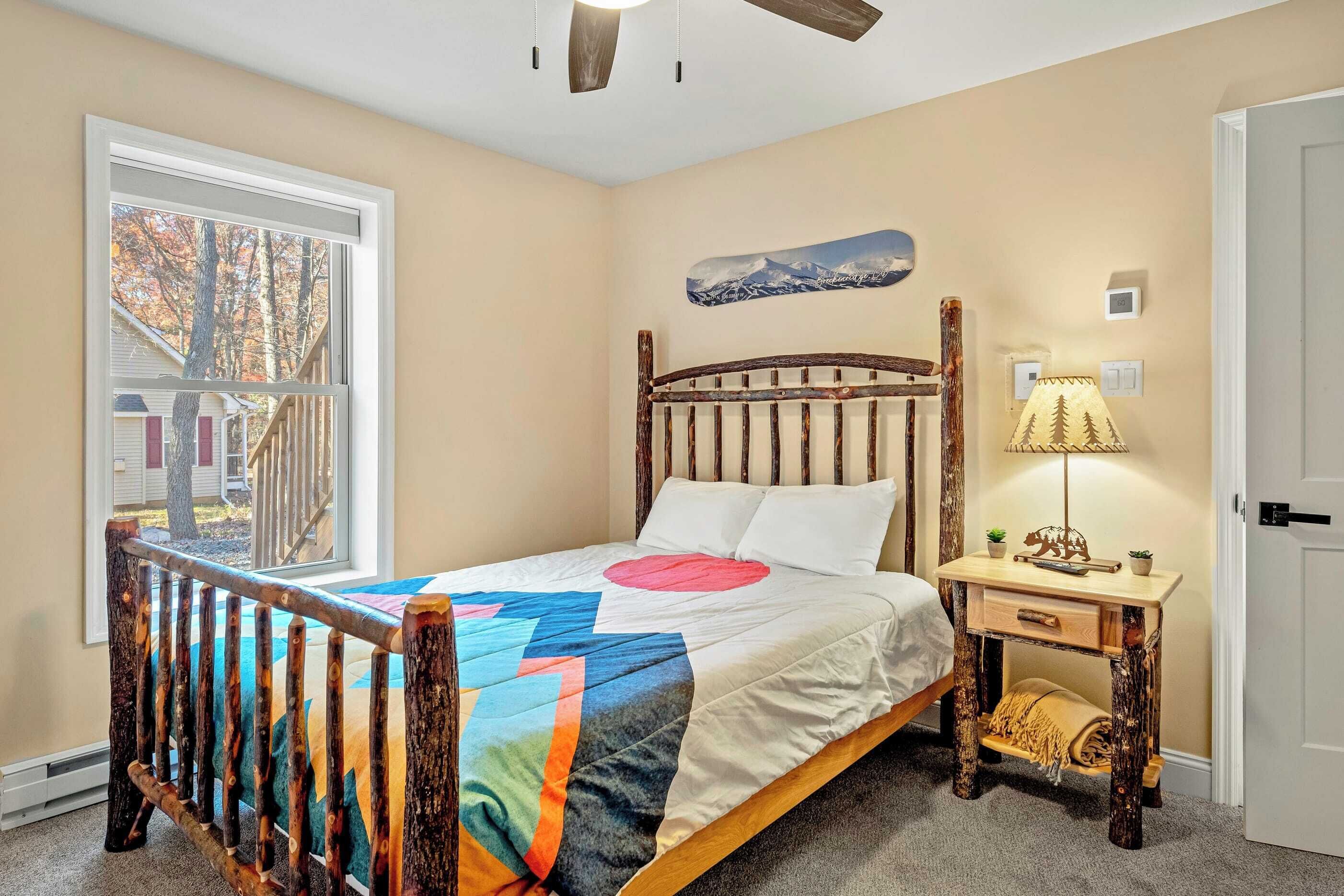 A bedroom with a wooden bed, a nightstand with a lamp, a window with a view of trees, and a wall-mounted picture.