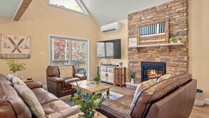 A cozy living room with leather sofas, a fireplace, a stone accent wall, and a large window overlooking a garden. - Stephen Way by Avantstay Hot Tub + Game Room Albrightsville (Albrightsville)
