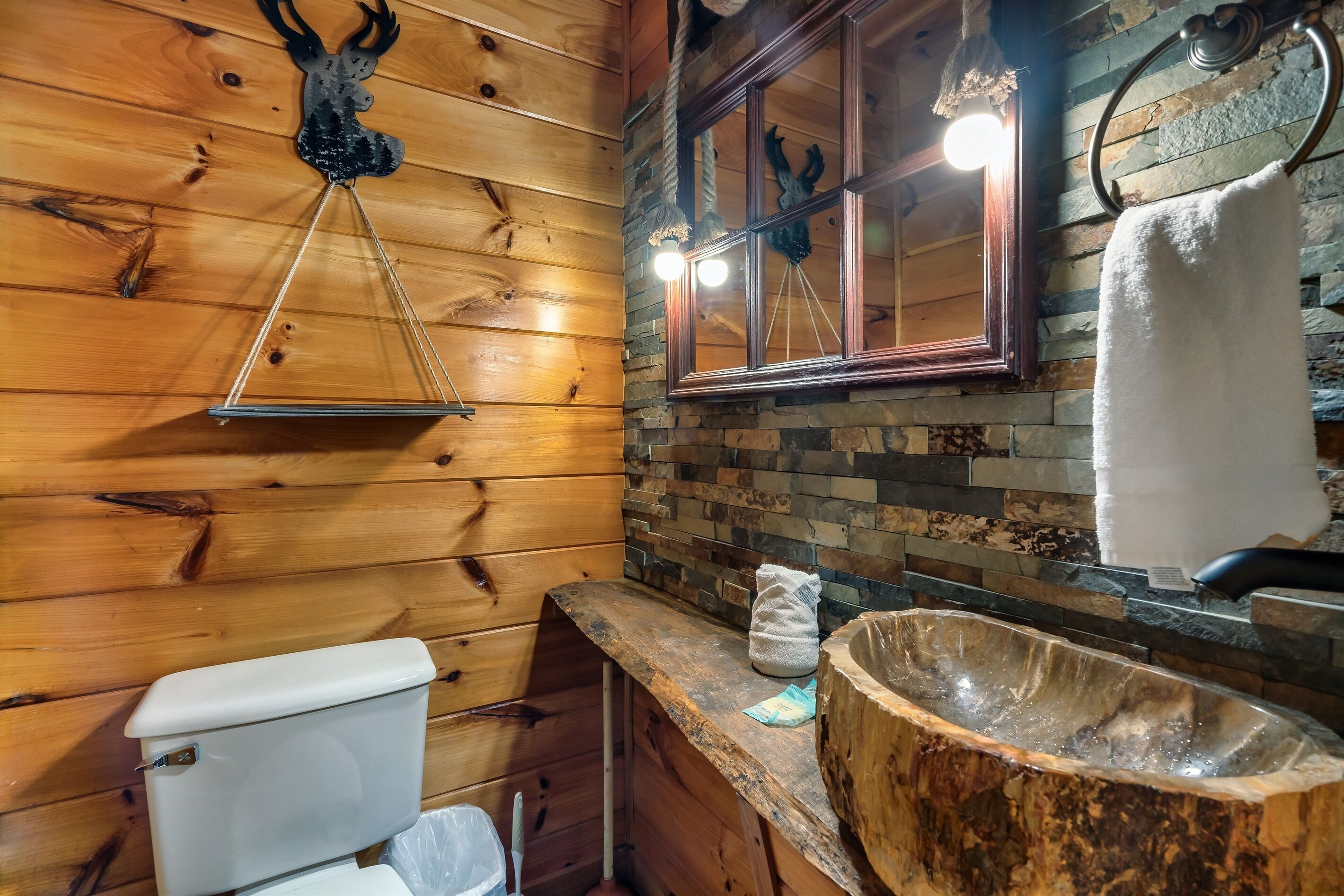 A rustic bathroom with wooden walls, a stone mosaic backsplash, a wooden sink basin, and a hanging deer head ornament.