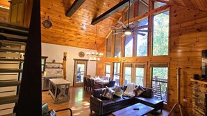 A wooden cabin interior with a dining area, sofa, fireplace, and large windows.