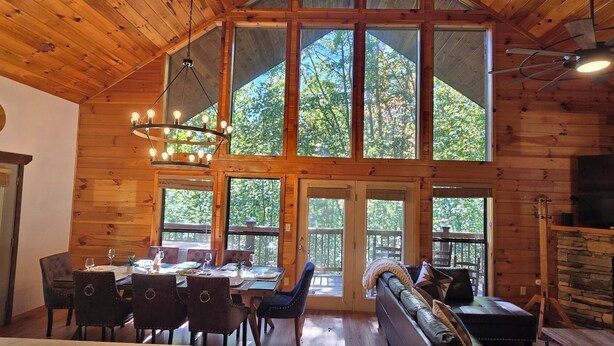 A wooden cabin interior with a dining area, large windows, a fireplace, and a ceiling fan.